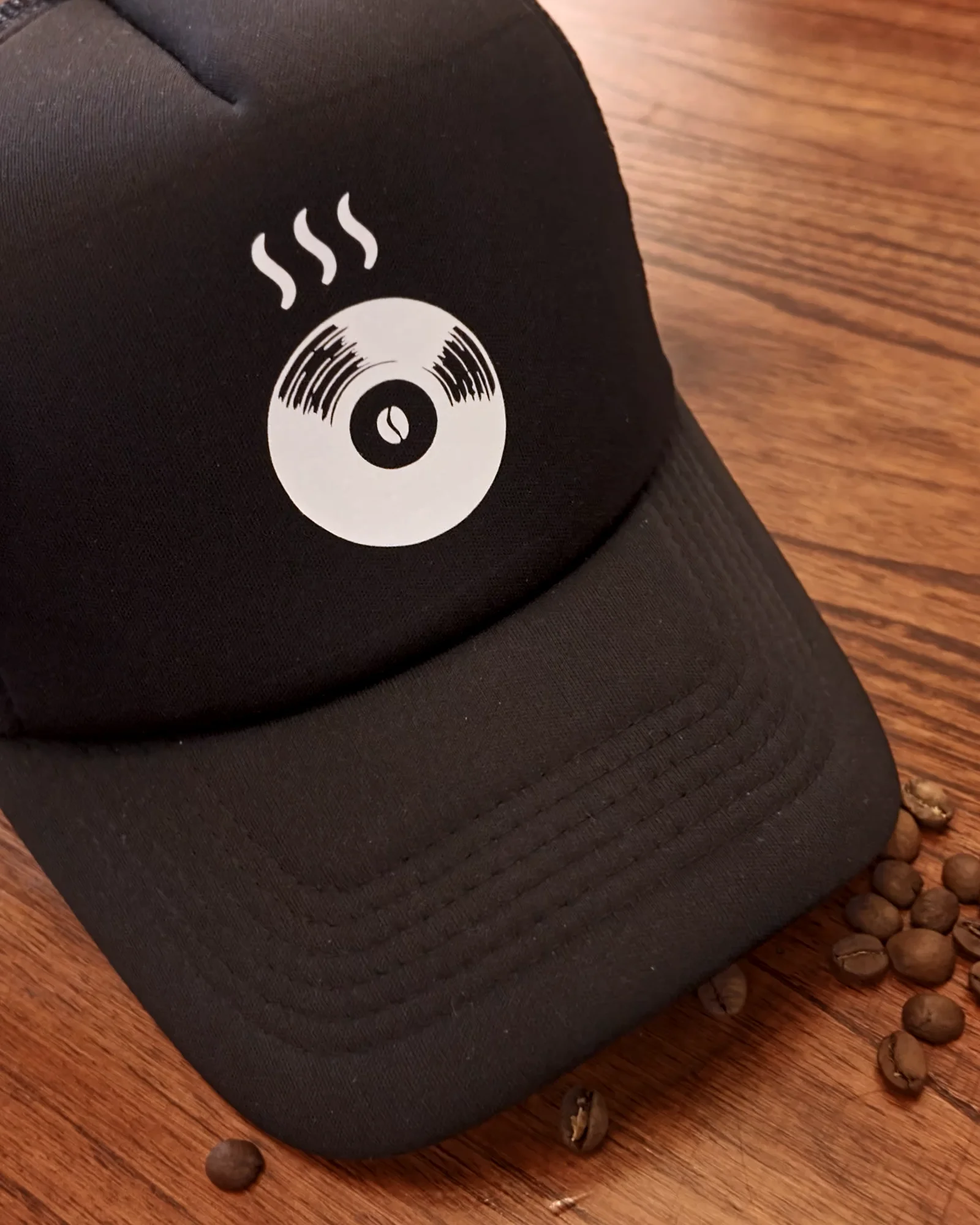Gorra trucker negra con el logo de Indómito en blanco, junto a un tamper y granos de café.